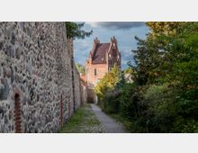 Pflasterweg zwischen einer hohen, grob gemauerten Steinmauer links und dichter Vegetation rechts, im Hintergrund ein historisches Backsteingebäude mit spitzen Giebeln und roten Dachziegeln.