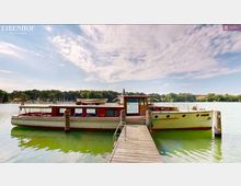Ein hölzernes Boot liegt an einem Steg auf einem ruhigen See. Im Hintergrund ist bewaldetes Ufer mit einigen Segelbooten und vereinzelten Gebäuden zu sehen, der Himmel ist leicht bewölkt.