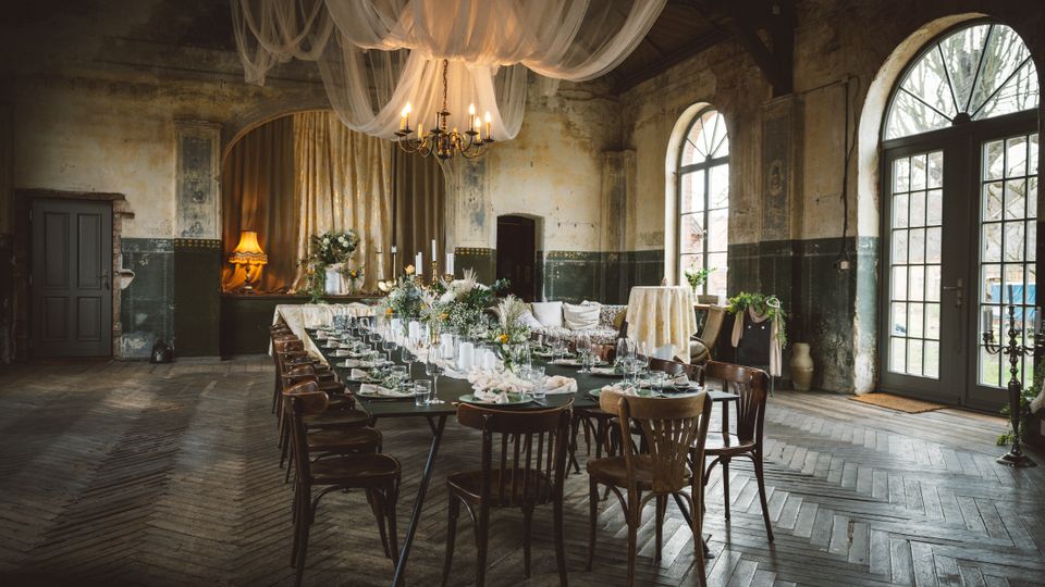 Rustikaler Festsaal in einem alten Industrie- oder Gutshausgebäude mit hohen Decken, abgenutzten Wänden und Holzparkett im Fischgrätmuster. In der Mitte steht ein lang gedeckter Tisch mit Blumenarrangements, darüber hängen ein Kronleuchter und drapierte Stoffbahnen; große Rundbogenfenster und Glastüren lassen Tageslicht herein.