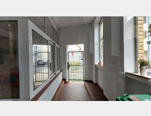 Überdachter Hausdurchgang mit gläsernen Fenstern und einer weißen Wand, durch den ein schmiedeeisernes Tor den Blick auf eine gepflasterte Straße und parkende Autos freigibt. Auf den Fenstersimsen stehen dekorative Blumentöpfe.