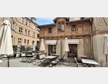 Innenhof mit Kopfsteinpflaster und einer Café-Terrasse mit Metalltischen, Stühlen und geschlossenen Sonnenschirmen. Im Hintergrund steht ein historisches Fachwerkgebäude mit großen Fenstern und Markisen, flankiert von weiteren Gebäudefassaden, Blumenbeeten und einer kleinen Statue.