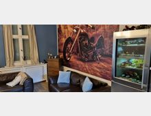 Wohnzimmer mit blauen Wänden, einem auffälligen Wandbild eines Motorrads, zwei braunen Ledersofas mit Kissen, einem weißen Fenster mit beigen Vorhängen und einer Vitrine mit Weinen. An der Wand steht zudem eine kleine Holzkommode mit Dekorationsgegenständen.