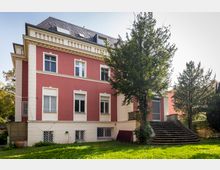 Rotes, zweistöckiges Wohngebäude mit weißen Fensterrahmen und Dachgauben, umgeben von einem Garten mit Rasen und Bäumen; auf der rechten Seite führen Steinstufen zu einem Eingang auf der erhöhten Terrasse.