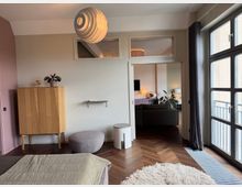 Bright modern apartment bedroom with herringbone wood floors, a large multi-pane window with dark curtains, and a sculptural pendant light. A wooden cabinet, round pouf, small side table, and a doorway with transom windows open into a living area beyond.