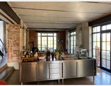 Offene Küche in einem Loft mit freigelegten Ziegelwänden, großen Fenstern mit schwarzen Rahmen und einer zentralen Kücheninsel aus Edelstahl. Im Hintergrund sind eine Treppe, Küchenutensilien und ein Pizzaofen sichtbar.