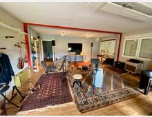 Aufnahmestudio mit hellem Holzboden, bunten Teppichen und mehreren Instrumenten, darunter Gitarren an der Wand, ein Schlagzeug und Verstärker. Im Hintergrund stehen ein großer Bildschirm, Lautsprecher und ein Tisch vor Fenstern mit Jalousien.