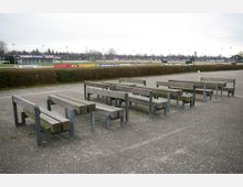 Außenbereich eines Sportplatzes mit einer Reihe von einfachen Holzbänken auf Metallgestellen auf einer gepflasterten Fläche. Im Hintergrund sind eine Hecke, Werbebanner und die mit Flutlichtmasten ausgestattete Spielfläche zu sehen.
