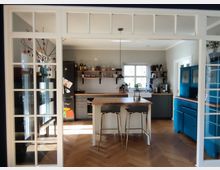 Moderne Küche mit grauen Unterschränken und einer Holzarbeitsplatte, zentral eine Kücheninsel mit zwei Barhockern. Links ein Kühlschrank mit Magneten, hinten ein Fenster mit Tageslicht, rechts ein blauer Schrank und ein Spülbereich mit weißem Waschbecken.