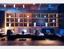 Eine moderne Lounge mit gepolsterten Sitzbänken und kleinen Tischen, umgeben von einem großen Regal mit dekorativen Flaschen und weißen Vasen. Die Beleuchtung betont die dunklen Holz- und Metallakzente, während blaue und graue Kissen die Sitzbereiche schmücken.