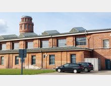 Backstein-Gebäude mit großen Fenstern und einem runden Turm im Hintergrund. Auf dem Vorplatz stehen mehrere geparkte Autos, und es sind moderne Anbauten wie Glasdächer auf dem Dach sichtbar.