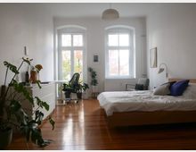 Helles Schlafzimmer mit Holzfußboden, einem großen Bett auf der rechten Seite, einem Schreibtisch mit Stuhl vor zwei großen Fenstern im Hintergrund und mehreren Zimmerpflanzen verteilt im Raum. Die Wände sind weiß, und die Möbel sind schlicht und modern gehalten.