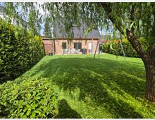 Großer, gepflegter Garten mit Rasenfläche, umgeben von hohen Hecken und überhängenden Zweigen eines Baumes. Im Hintergrund steht ein Backsteinhaus mit Terrasse und Sitzmöbeln; rechts befindet sich ein Schaukelgestell.