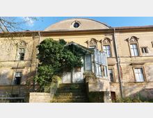 Eine verfallene historische Villa mit gelblicher Fassade und dekorativen Fensterumrahmungen. Im Vordergrund führt eine mit Moos bewachsene Treppe zu einem verwitterten, überwucherten Wintergarten mit Glastüren.