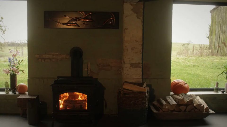 Ein rustikaler Innenraum mit einem schwarzen Kaminofen, in dem Feuer brennt, umgeben von einer grünen Wand mit sichtbaren Ziegeln. Auf beiden Seiten befinden sich große Fenster mit Blick ins Freie, Holzscheite in einem Korb und herbstliche Dekoration wie Kürbisse und Blumen.