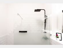 Moderne Dusche mit schwarz-mattem Duschkopf und Glastrennwand in einem Badezimmer mit weißen Wandfliesen. Auf der Fensterbank steht eine kleine Pflanze in einem weißen Blumentopf.