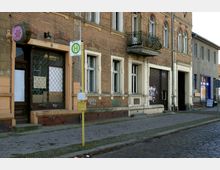 Straßenszene mit mehrstöckigem Gebäude aus Backstein, das heruntergekommene Fassaden und Graffiti aufweist. Eine Bushaltestelle mit einem gelben Schild steht vor dem Gebäude, daneben ein Geschäft mit Gitterschaufenster und geschlossenem Rolltor.