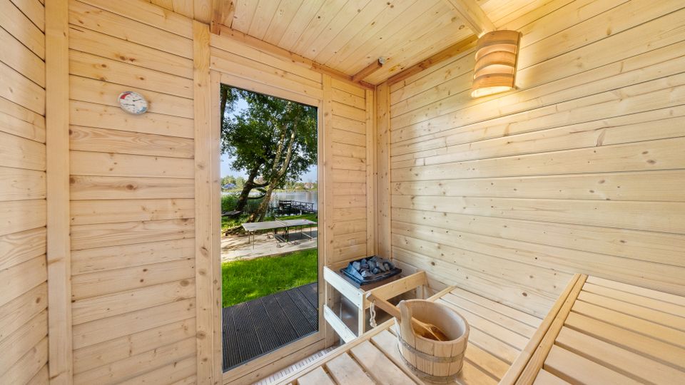Innenraum einer kleinen Holzsauna mit hellen Holzvertäfelungen, Saunabänken, Saunaofen mit Steinen, Eimer und Thermometer. Durch die Glastür blickt man auf eine Grünfläche mit Baum und Sitzgelegenheit nahe einem Gewässer.