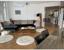 Wohnzimmer in einer modernen Wohnung mit hellen Holzböden, einer großen Ecksofa-Garnitur mit Kissen und einem Couchtisch aus Metall und Glas. Im Vordergrund steht ein Esstisch mit einer Holzoberfläche, im Hintergrund ist eine offene Tür zur Küche erkennbar; dekorative Beleuchtung und Teppiche runden die Einrichtung ab.