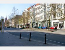 Straßenszene in einer Stadt mit mehrstöckigen Häusern im Gründerzeitstil, Geschäften im Erdgeschoss und geparkten Autos entlang der Straße. Im Hintergrund ragt der Turm einer Kirche in den Himmel, kahle Bäume säumen die Straße.