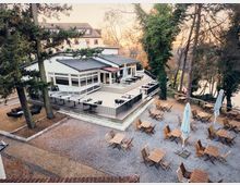 Outdoor dining area surrounded by tall trees, featuring wooden tables and chairs arranged on a gravel surface, alongside a modern white building with large windows and a raised terrace. The setting is within a wooded landscape, with warm light suggesting a sunset in the background.