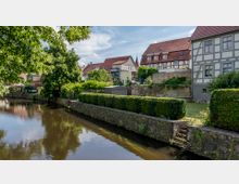 Fachwerkhäuser mit roten Ziegeldächern säumen einen ruhigen Flusslauf, der von einer Steinmauer und gepflegten Hecken begrenzt ist. Im Vordergrund stehen Bäume und Grünflächen, während im Hintergrund eine kleine dörfliche Umgebung sichtbar ist.