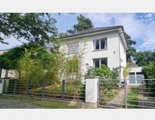 Zweistöckiges weißes Einfamilienhaus mit flachem Dach und großen Fenstern, umgeben von Bäumen und Bambuspflanzen. Im Vordergrund befindet sich ein Metallzaun mit einem Gehweg, der zu einer Garage rechts neben dem Haus führt.