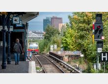 Bahnhofsszene mit einem einfahrenden S-Bahn-Zug auf einem eingleisigen Gleis, umgeben von Bäumen und Graffiti-bedeckten Wänden. Im Hintergrund sind moderne Hochhäuser und eine Brücke zu sehen, während ein Fußgänger den Bahnsteig entlangläuft.