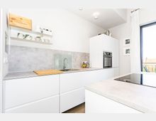 Helle, moderne Küche mit weißen Schränken, grauer Arbeitsplatte und Rückwand in Betonoptik. Die Ausstattung umfasst einen eingebauten Backofen, ein Glaskeramik-Kochfeld, eine Spüle mit Armatur und offene Regale mit Geschirr; ein Fenster mit Vorhängen sorgt für Tageslicht.