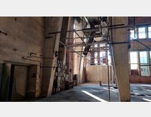 Industriehalle eines verlassenen Fabrikgebäudes mit massiven Betonstützen, großen Fenstern mit teils zerbrochenem Glas und verwitterten Wänden. Im Raum befinden sich eine alte Schalttafel mit Kabeln und verlassene Metallgerüste unter der Decke.