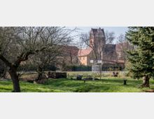Grüner Garten mit Rasenflächen und einzelnen Bäumen im Vordergrund, dahinter ein Sportplatz mit Basketballkörben und ein Gebäudeensemble, dessen markanter Kirchturm aus Backstein hervorragt. Im Hintergrund stehen weitere Bäume und Gebäude.