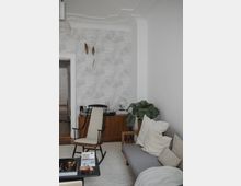 Wohnzimmer mit heller, moderner Einrichtung, bestehend aus einem grauen Sofa mit Kissen, einem schwarzen Schaukelstuhl und einem niedrigen Couchtisch. Im Hintergrund stehen ein Holz-Sideboard mit Deko-Elementen und eine große Zimmerpflanze neben einer Tür, die in den Flur führt. Wände sind in hellem Grau gestaltet, teils mit einer gemusterten Tapete.