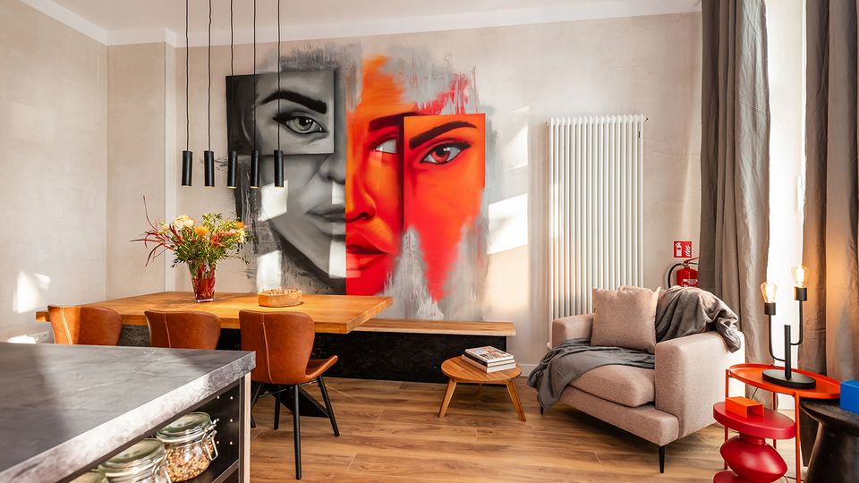 Moderner Wohn- und Essbereich mit Holztisch und Lederstühlen, schwarzer Pendelleuchte sowie einer Sitzecke mit Sessel, kleinem Couchtisch und Beistelltisch. An der Wand hängen großformatige, farbige Porträtgemälde; daneben stehen ein hoher weißer Heizkörper und ein bodentiefes Fenster mit grauen Vorhängen.