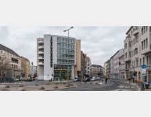 Straßenszene in einer Stadt mit mehrstöckigen Wohn- und Geschäftshäusern. Im Vordergrund befindet sich eine Kreuzung mit Verkehrsinseln und Ampeln, die von umliegenden Gebäuden in unterschiedlichen Architekturstilen eingerahmt wird.