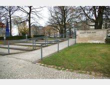 Eine gepflasterte Rampe mit Metallgeländern führt durch eine Grünfläche mit Bäumen auf den Theodor-Heuss-Platz. Im Hintergrund sind Gebäude und ein blauer Obelisk zu sehen.