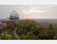 Ehemalige Abhörstation mit geodätischer Kuppel auf einem Betongebäude, umgeben von herbstlich gefärbtem Wald und einem grauen, bewölkten Himmel. Im Vordergrund sind Gebäudereste mit Graffiti sowie eine schmale asphaltierte Straße zu sehen.