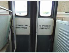 Innenansicht einer Bustür mit zwei kleinen Fenstern; auf den Türflächen befinden sich weiße Schilder mit der Aufschrift „TO OPEN DOOR PUSH HANDLE“. Links ist ein Metallhandlauf zu sehen, rechts eine Wandverkleidung mit geriffeltem Muster.