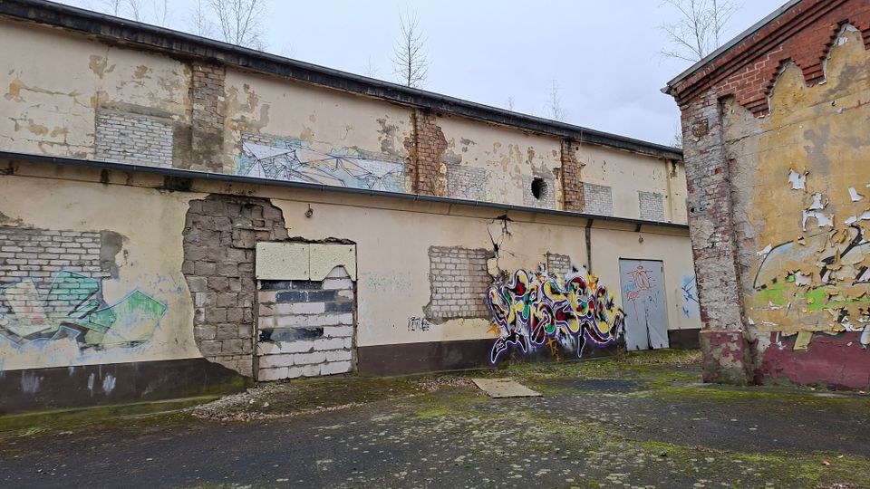 Verwitterter Hinterhof mit einer langen, flachen Gebäudefassade aus bröckelndem Putz und freiliegendem Mauerwerk, teils zugemauerten Öffnungen und Graffiti. Rechts steht ein weiteres Gebäude mit abplatzender Farbe; der Boden wirkt rissig und moosbewachsen.