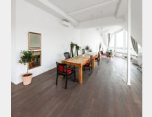 Heller, modern eingerichteter Raum mit dunklem Holzboden, weiß gestrichenen Wänden und großen Fenstern, die viel Tageslicht hereinlassen. Ein langer Holztisch mit verschiedenen Stühlen steht im Vordergrund, mit Pflanzen und Dekorelementen verteilt im Raum.