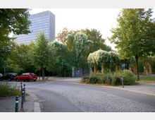 Ein gepflasterter Weg in einer parkähnlichen Umgebung mit vereinzelten Bäumen, einer begrünten Pergola und einem modernen Hochhaus im Hintergrund. Am Straßenrand stehen parkende Autos, und eine Person ist in der Nähe der Pergola zu sehen.
