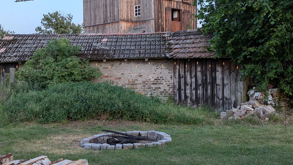Alte hölzerne Windmühle mit Flügeln und Schindeldach hinter einem niedrigen Nebengebäude auf einem ländlichen Grundstück. Im Vordergrund liegen gestapelte Brennholzscheite, daneben eine steinerne Feuerstelle, umgeben von Wiese und Bäumen.
