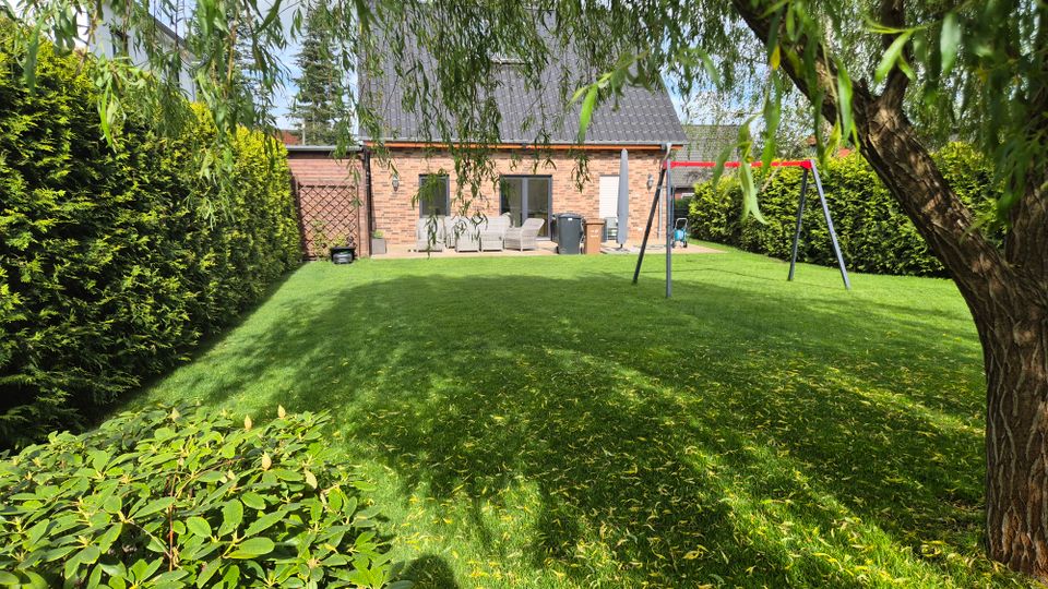 Großer, gepflegter Garten mit Rasenfläche, umgeben von hohen Hecken und überhängenden Zweigen eines Baumes. Im Hintergrund steht ein Backsteinhaus mit Terrasse und Sitzmöbeln; rechts befindet sich ein Schaukelgestell.