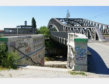 Brücke aus Metallstreben im urbanen Raum mit einer graffitibedeckten Treppenunterführung im Vordergrund. Im Hintergrund ist ein modernes Gebäude mit einem "S-Bahn"-Zeichen und ein Baum zu sehen.