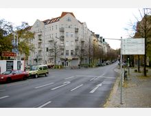 Straßenkreuzung in einer städtischen Umgebung mit mehrstöckigen Wohnhäusern im klassischen Baustil, einigen Balkonen und roten Dachziegeln. Im Vordergrund sind parkende Autos, ein Straßenschild mit Routenangaben zu nahegelegenen Zielen und ein Gehweg mit kahlen Bäumen sichtbar.