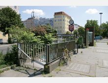 Ein Straßenzug mit einer Eingangstreppe zu einer öffentlichen Toilette, gekennzeichnet durch ein Schild mit der Aufschrift "HERREN". Im Vordergrund steht ein gelbes Fahrrad angelehnt an ein Geländer, im Hintergrund sind Bäume und Gebäude mit unterschiedlichen Fassaden sowie eine Uhr und Werbeschilder sichtbar.