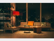 Gemütlich eingerichteter Loungebereich mit einer beigefarbenen Polstercouch, dekorativen Kissen, einem kleinen Couchtisch und einer roten Stehlampe. Im Hintergrund sind große Glasfenster sowie ein Regal mit Gläsern zu sehen, beleuchtet von warmem Licht.