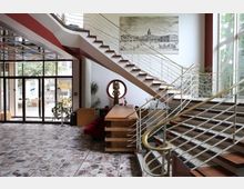 Foyer eines modernen Gebäudes mit einer großen Wendeltreppe und Metallgeländern, Marmorboden und einer Glasfront mit Blick auf einen Außenbereich. An der Wand hängt ein historisches Bild, daneben ein runder Spiegel; Pflanzen, eine Holztheke und rote Stühle ergänzen die Einrichtung.