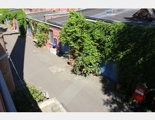 Innenhof mit asphaltierter Fläche neben einem flachen Backsteingebäude, dessen blaue Türen und Fenster von dichtem Efeu und Kletterpflanzen überwuchert sind. Mehrere Pflanzkübel stehen entlang der Wand, und ein rotes Schild markiert den Zugang im rechten Bildbereich.