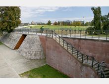 Treppen und Gehwege aus Sandstein führen über eine steinerne Rampe mit Geländern in einer parkähnlichen Umgebung mit Rasenflächen und Bäumen. Im Hintergrund sind moderne Gebäude und der Berliner Fernsehturm sichtbar.