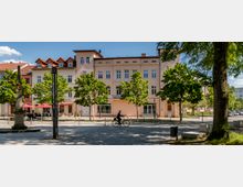 Straßenszene mit einem Platz, gesäumt von Bäumen und einer Reihe von pastellfarbenen, mehrstöckigen Gebäuden. Im Vordergrund fährt eine Person auf einem Fahrrad über das Kopfsteinpflaster, daneben ein historischer Obelisk und Sitzbänke unter den Bäumen.