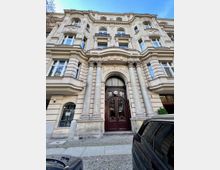 Mehrstöckiges historisches Gebäude mit beiger Fassade, verzierten Reliefs und Balkonen. Das Eingangsportal ist von zwei massiven Säulen flankiert und besitzt eine dunkle, hölzerne Doppeltür mit Glaselementen.